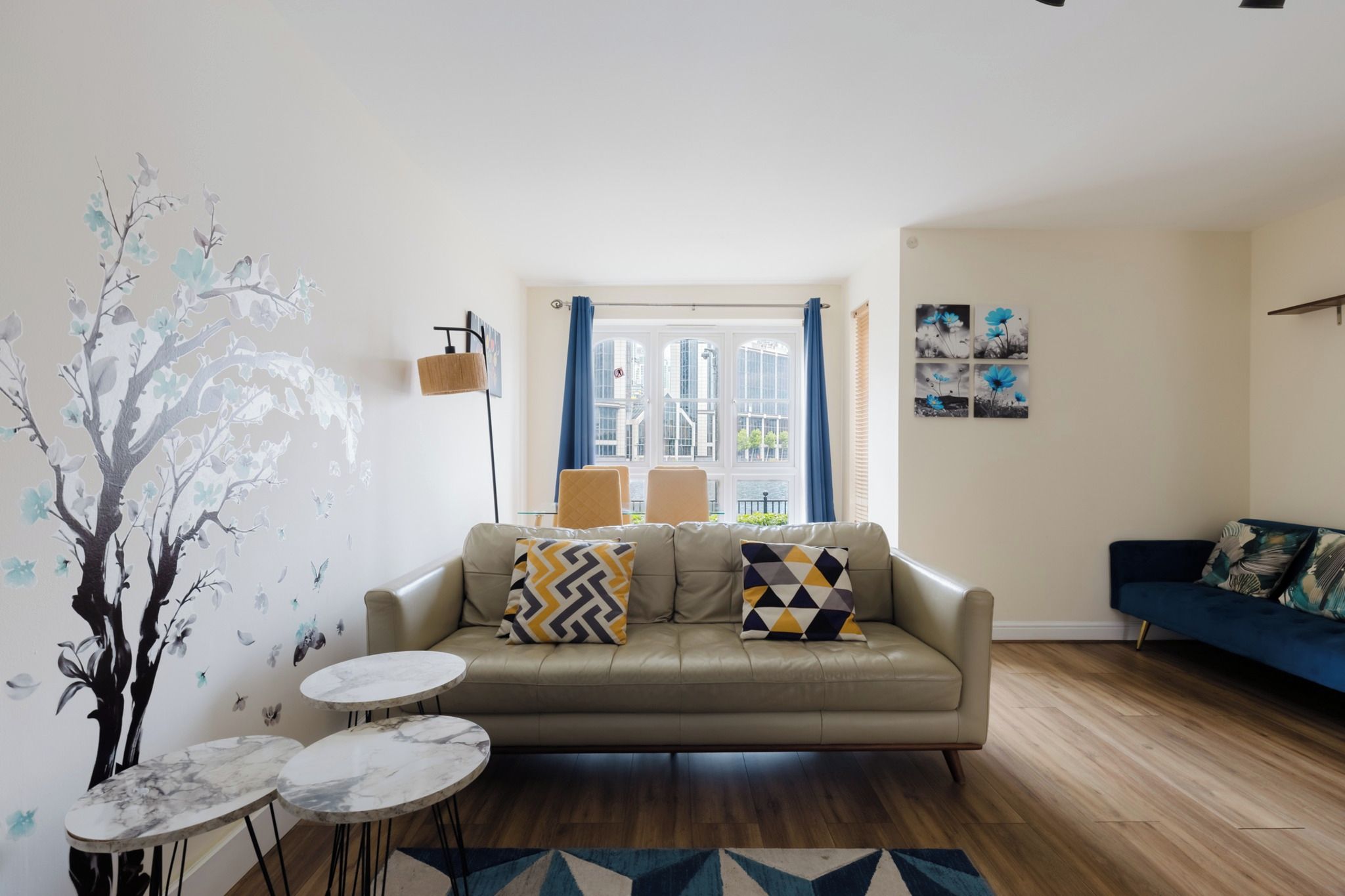 Una sala de estar con sofá, dos mesitas auxiliares, una lámpara y vistas a un paisaje urbano a través de amplios ventanales.