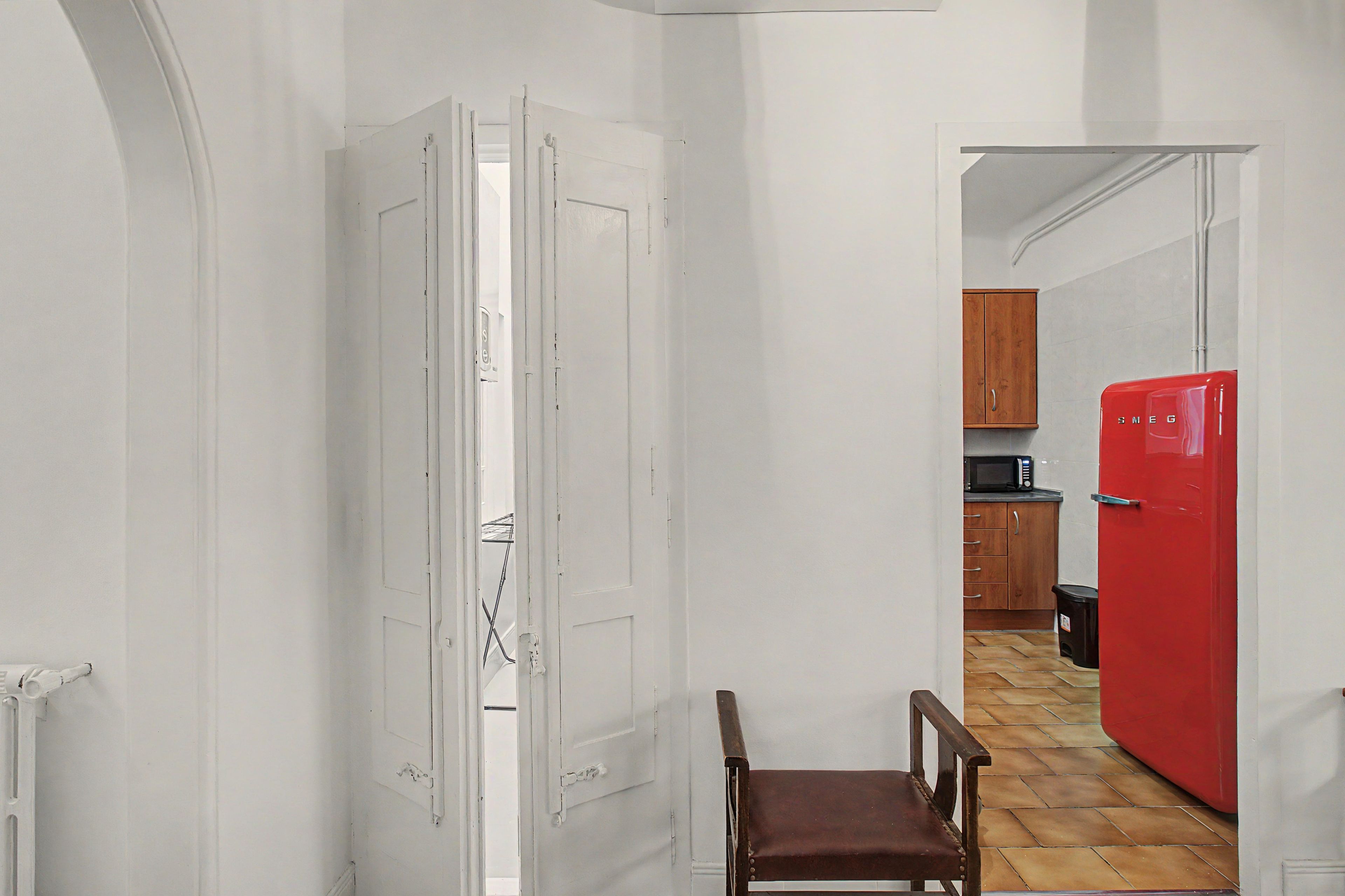 A narrow hallway with a white door, a wooden chair, and a red refrigerator in a kitchen area.