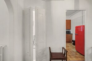 Un couloir étroit avec une porte blanche, une chaise en bois et un réfrigérateur rouge dans un coin cuisine.