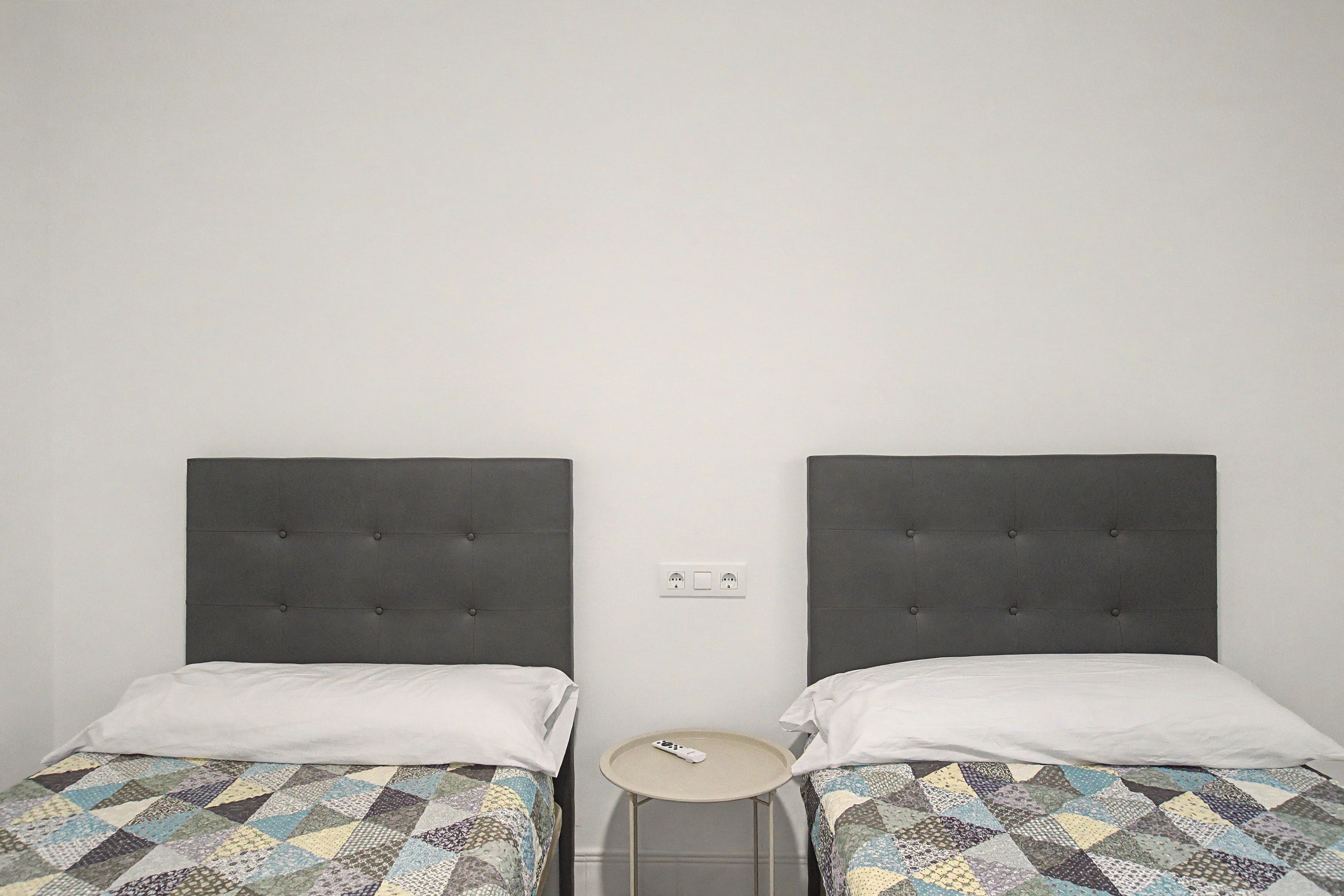 Dos camas individuales con cabeceras tapizadas en gris, una mesita redonda y un enchufe en la pared.
