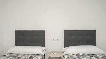 Dos camas individuales con cabeceras tapizadas en gris, una mesita redonda y un enchufe en la pared.