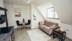 A modern living room with a sofa, a dining table with chairs, a kitchen area, and a large window. - Queensferry Collection - Residence 7 (Derby)