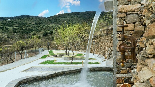 Ein Steinbrunnen mit Wasserfall, umgeben von einem angelegten Garten mit Wegen und Bäumen.