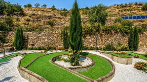Ein angelegter Garten mit Steinmauern, einer zentralen, runden Rasenfläche und einer Reihe von Solarmodulen im Hintergrund.