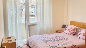 Ett sovrum med en säng, ett sängbord, en lampa, ett fönster med gardiner och en dörr.