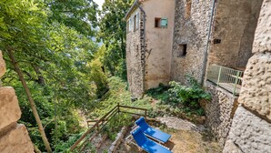 Un bâtiment en pierre doté d’une petite fenêtre et d’un escalier en bois menant à un espace extérieur aménagé avec des sièges bleus.