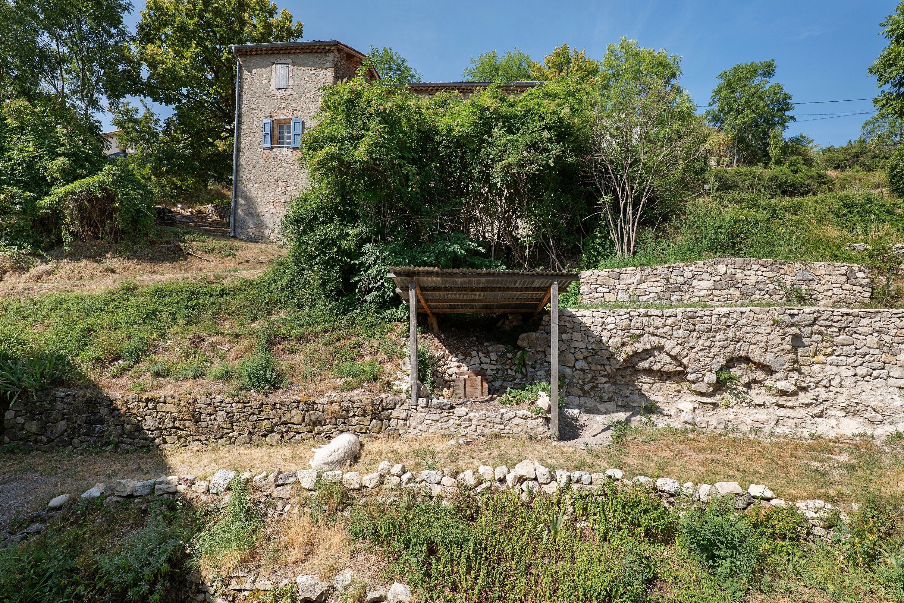 Un edificio de piedra con una estructura de madera en primer plano, rodeado de follaje y árboles.