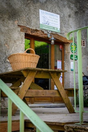 Eine rustikale Holzbank mit einem Korb aus Weide, ein Steinhaus mit dem Schild 'LA CHAMBRETTÉ' und ein grüner Metallzaun.