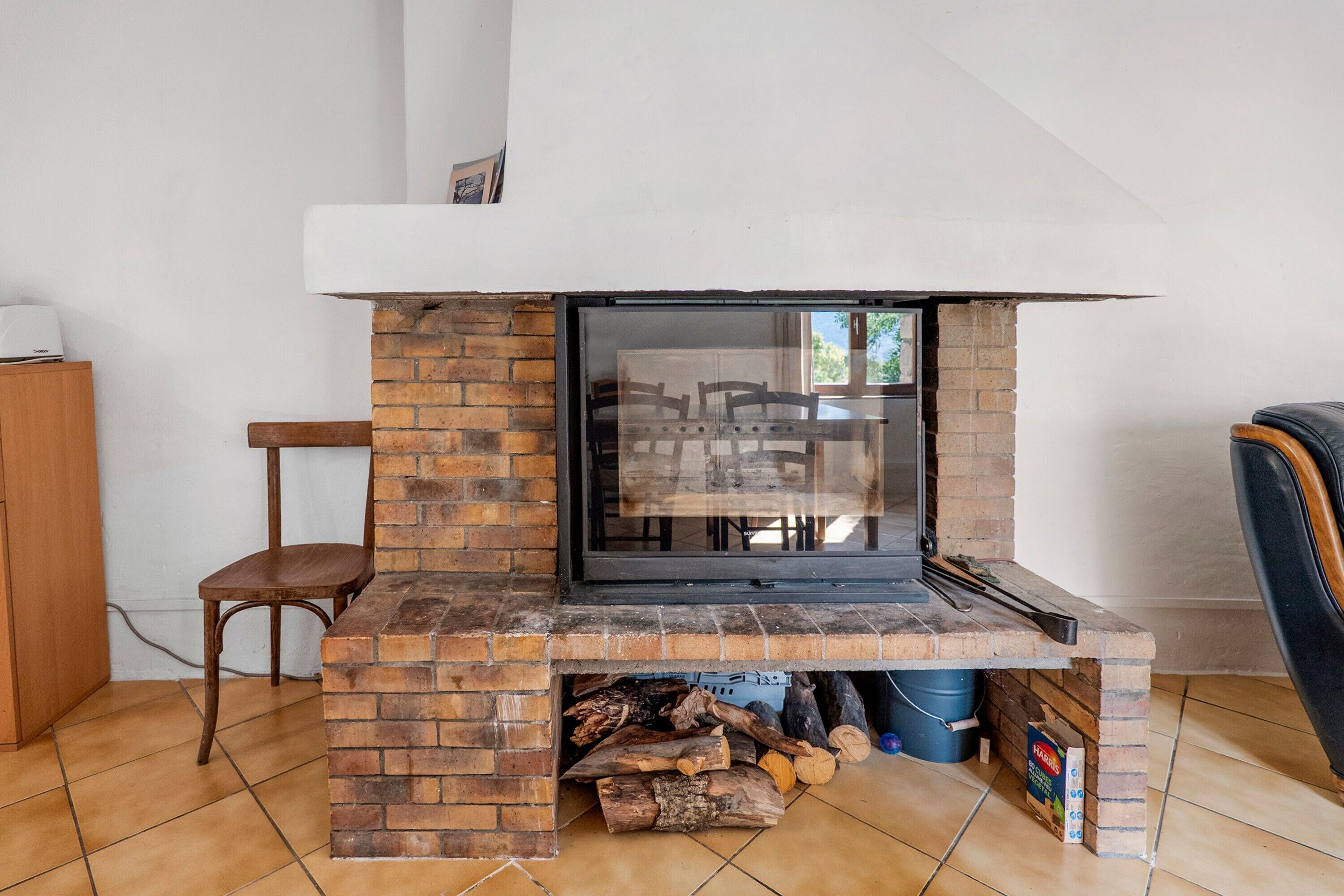 Een metselwerk open haard met een glazen deur, een houten stoel en een eettafel zichtbaar door het glas.