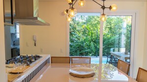 A kitchen with a gas stove, dining table, chairs, and a view of greenery outside. - Casa Serena | Oceanfront, Heated Pool, Jacuzzi & Fireplace (Florianópolis)