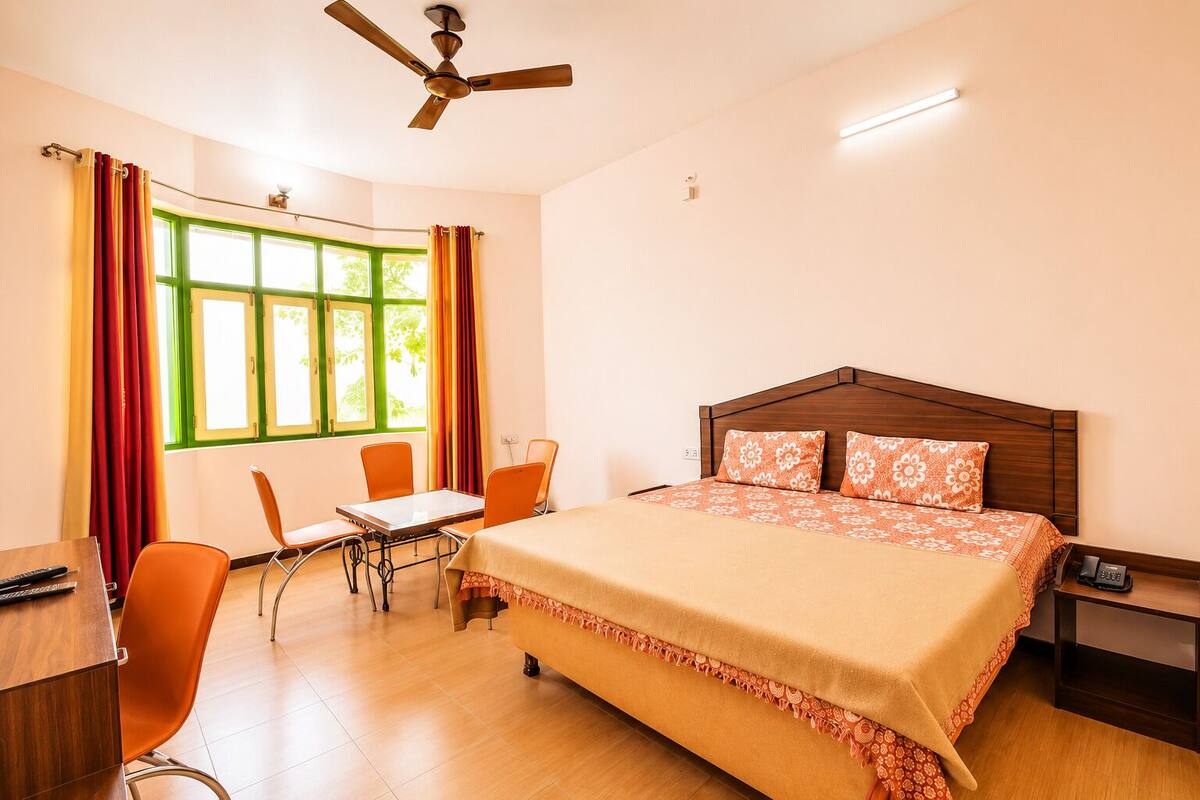 A hotel room with a bed, a desk, a chair, a table, a ceiling fan, and a window with curtains.