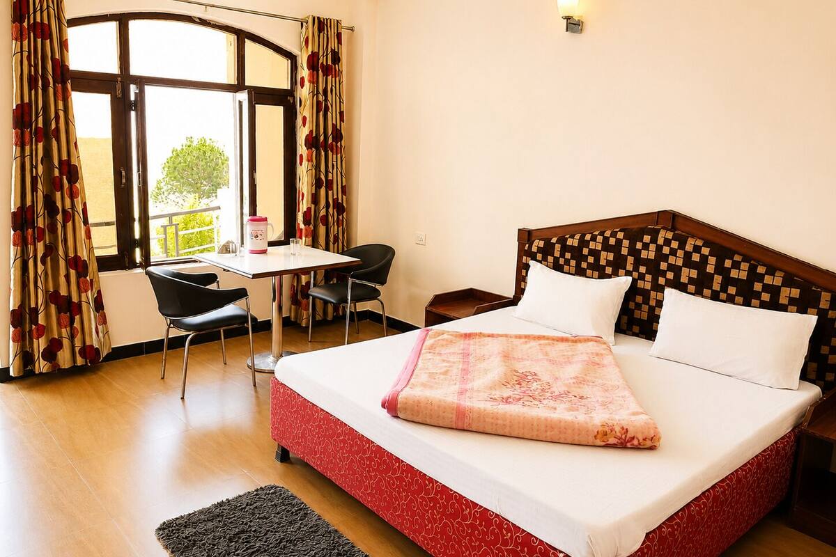 A hotel room with a bed, a desk, two chairs, a window with curtains, and a nightstand.