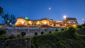 A multi-story hotel with a balcony on the second floor, a stone retaining wall, and a green sign indicating the hotel's name.