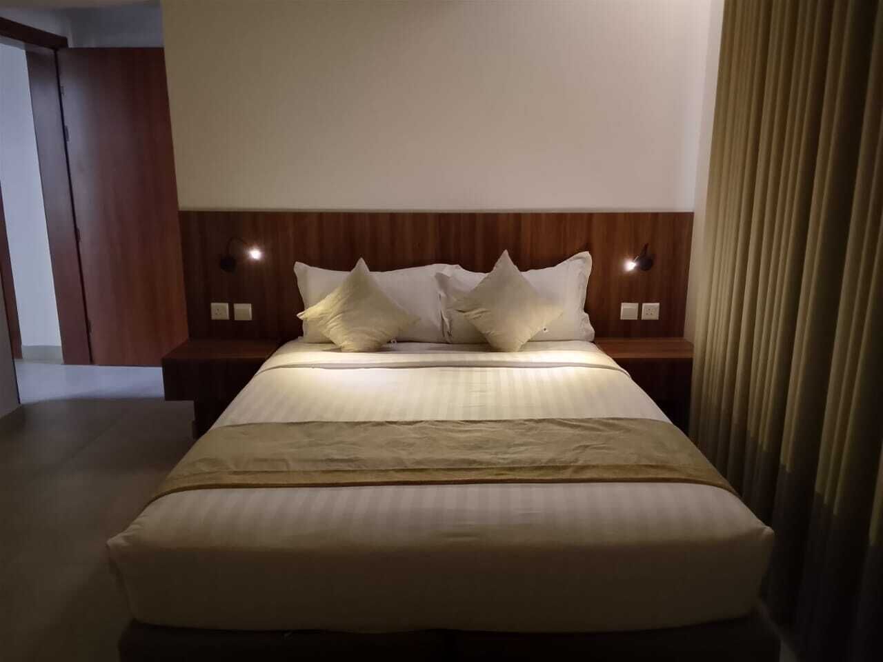 Habitación de hotel con una cama grande, cabecera de madera, dos mesitas de noche y una cortina a la derecha.