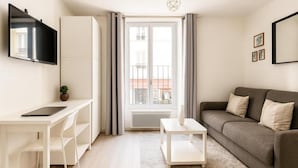 Una sala de estar moderna con un escritorio blanco, un sofá gris, un televisor de pantalla plana montado en la pared y un ventanal con cortinas.