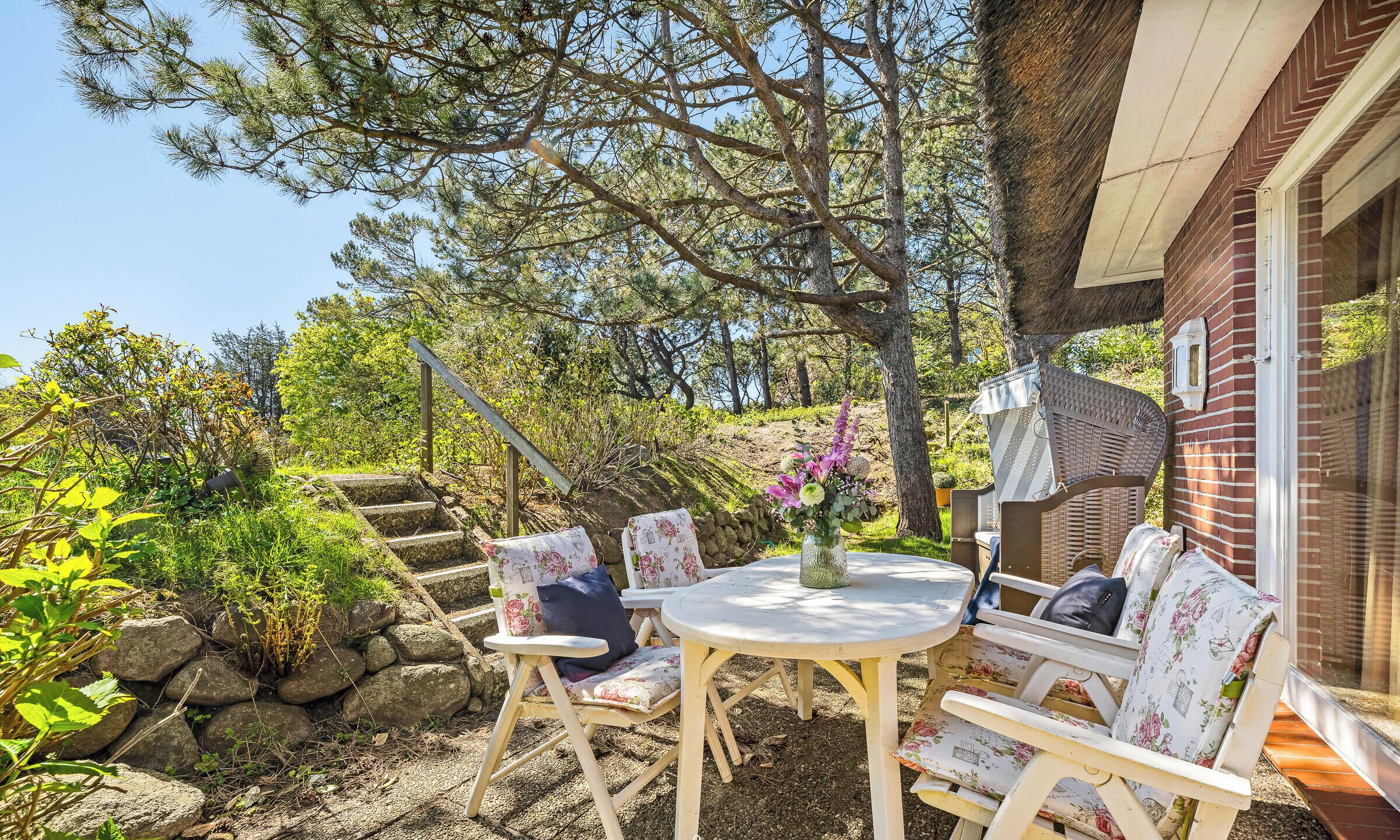 Een terras met een ronde tafel en witte stoelen, omringd door groen en een stenen muur.