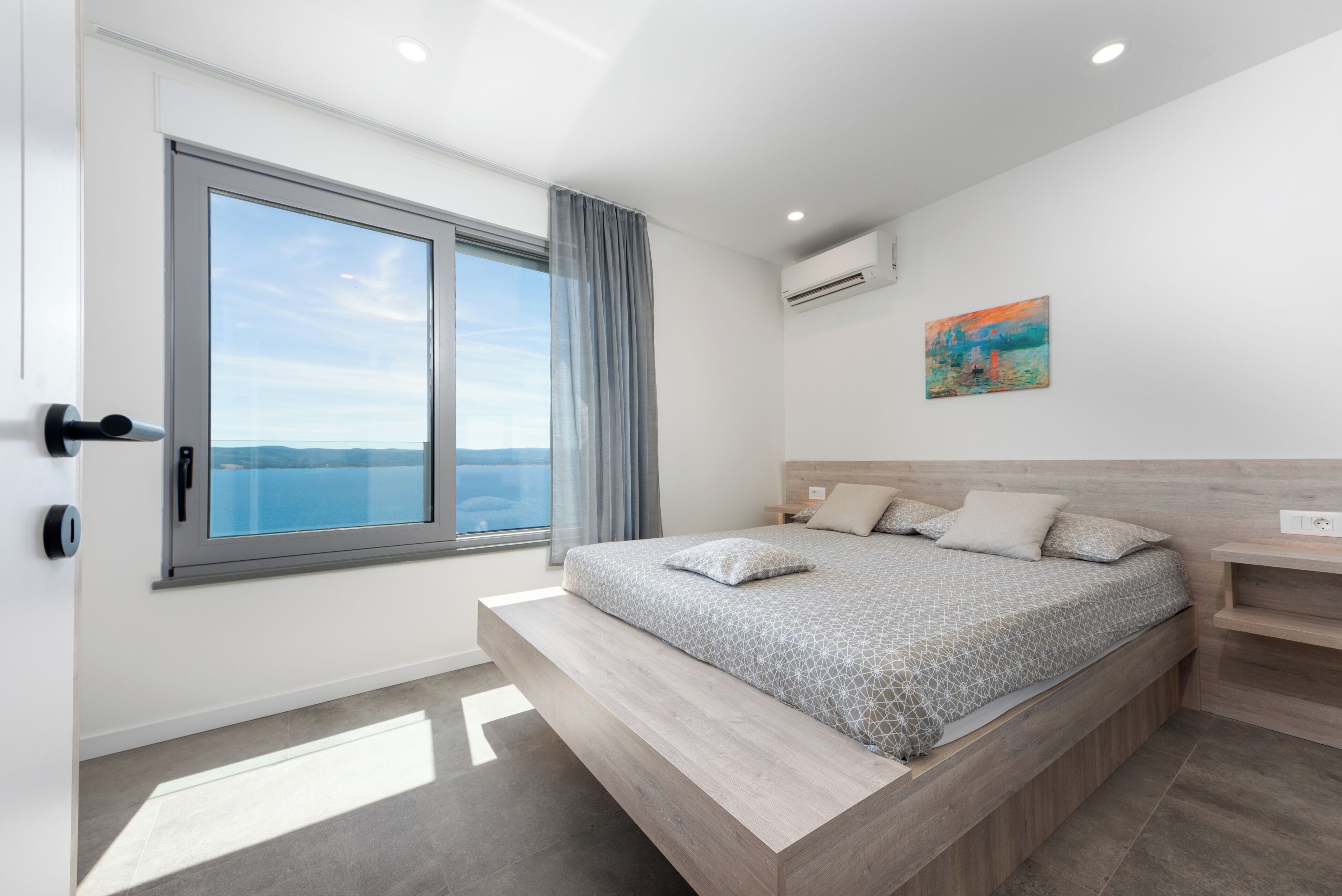 A modern bedroom with a large window, a bed with patterned bedding, a headboard, and a painting on the wall.
