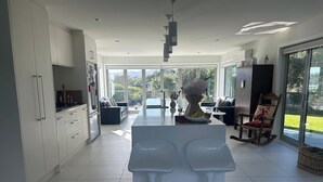 Een moderne keuken met witte kasten, een centraal eiland met barkrukken, een zithoek met een bank, en grote ramen met uitzicht op het buitengebied.