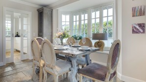 A dining area with a rustic wooden table, chairs, and a view of greenery through large windows. - Wääfblik – Your vacation home on Sylt with a sauna and ocean view (Wenningstedt-Braderup (Sylt))