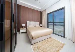 A modern bedroom with a large bed, bedside lamps, a nightstand, and a view of the beach through a large window. - Seafront Apt w/ Pool & Gym near Attractions (Sharjah)