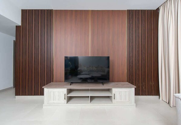 A modern living room with a flat-screen TV on a wooden entertainment unit, a large window with curtains, and a white floor.
