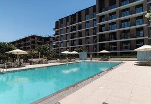 Une piscine entourée de chaises longues et de parasols, elle-même bordée d’un immeuble à plusieurs étages doté de balcons.