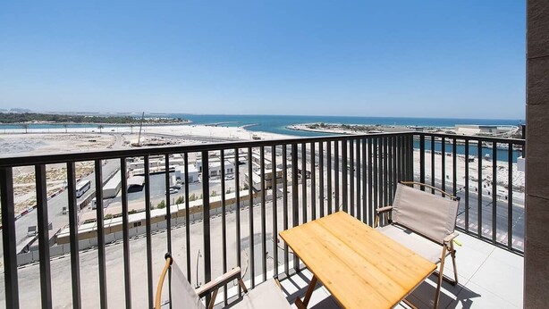 A balcony with a wooden table and chairs, overlooking a coastal area with a clear view of the sea and a sandy beach. - Seafront Apt w/ Pool & Gym near Attractions (Sharjah)
