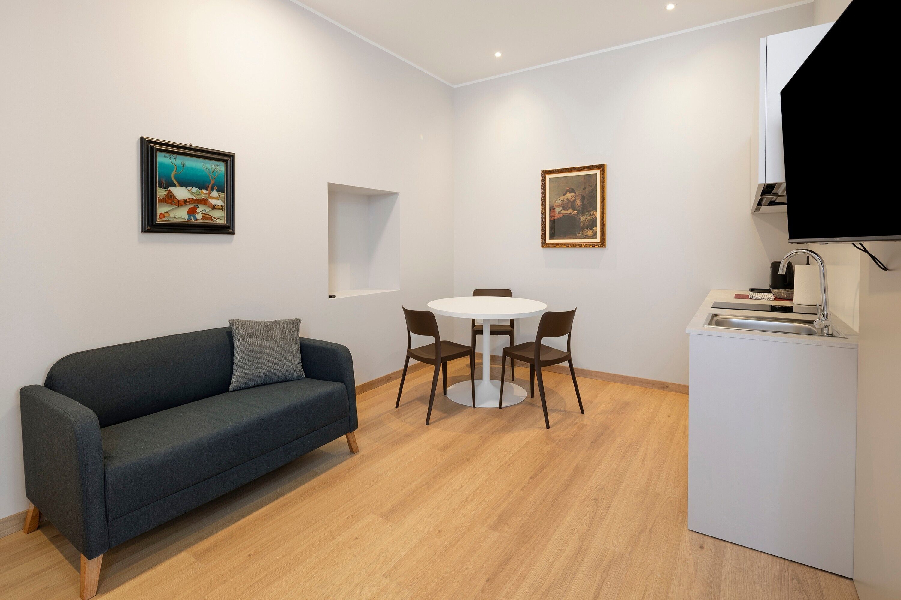 Habitació moderna amb sofà, taula rodona i cadires, zona cuina petita i dos quadres a les parets.