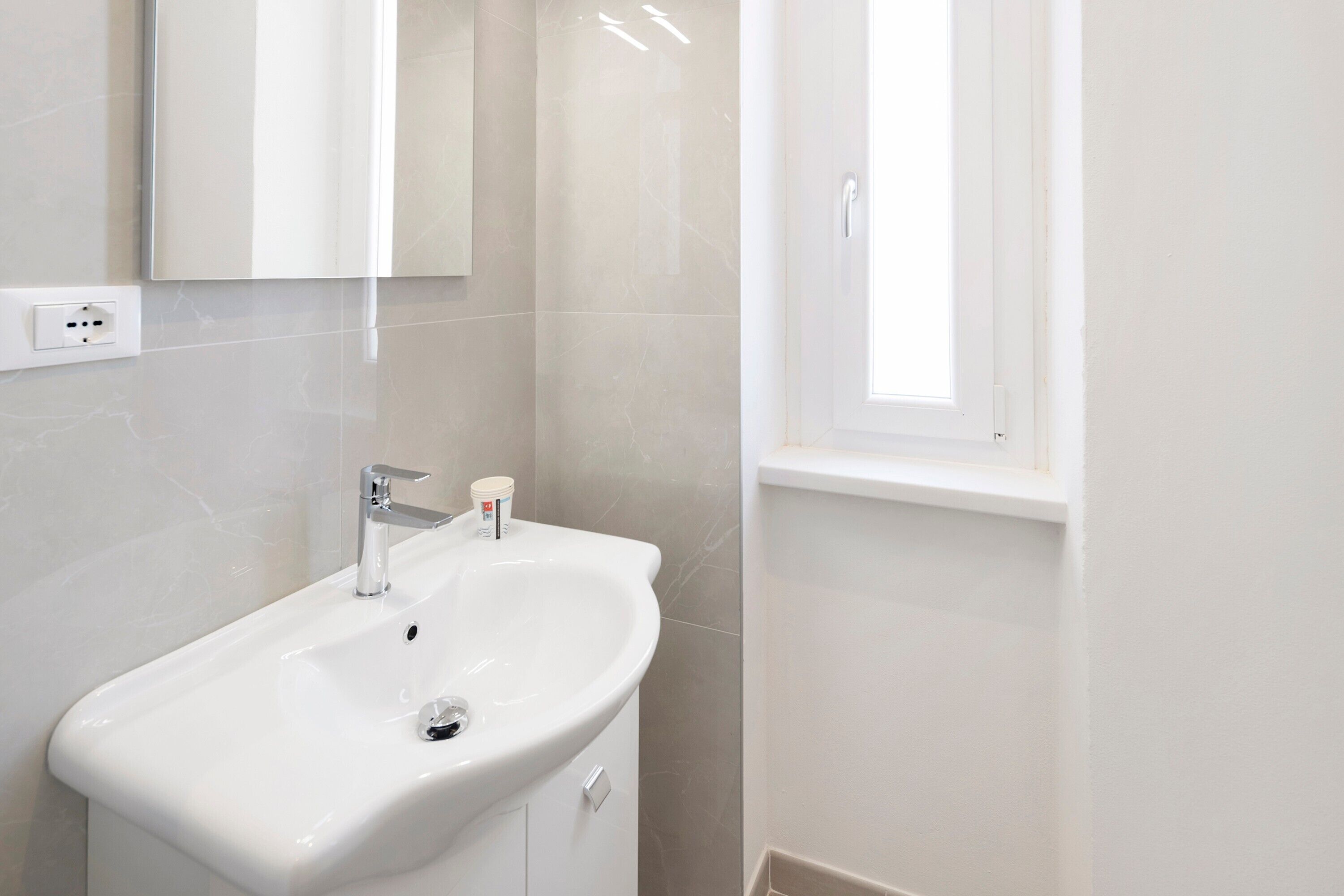 Een badkamer met een witte wastafel, spiegel en een raam.