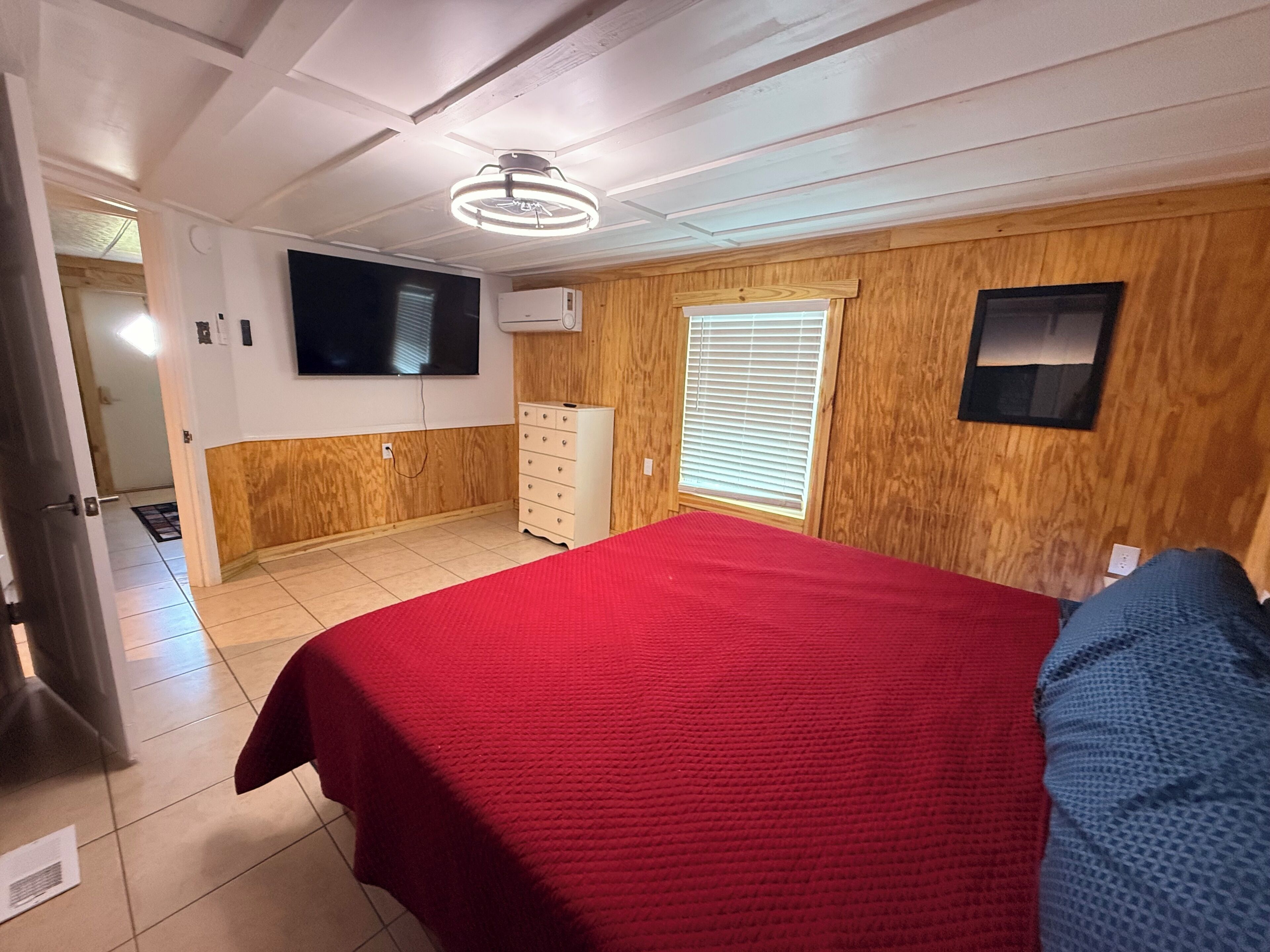 Une chambre avec une couvre-lit rouge, des murs lambrissés, un ventilateur de plafond, un téléviseur fixé au mur et une fenêtre avec des stores.