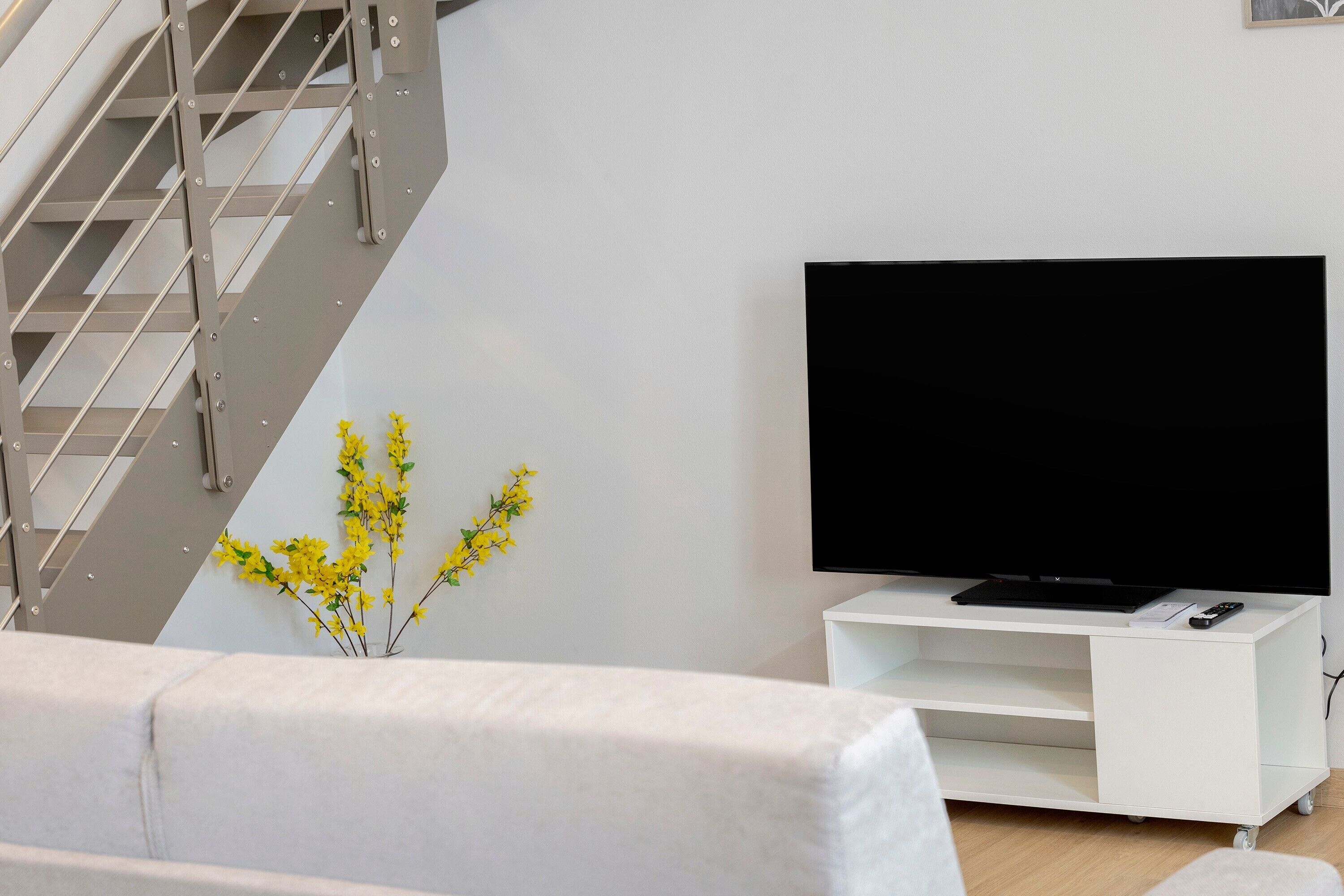 Un salon moderne avec un escalier, un téléviseur à écran plat posé sur un meuble blanc et une petite plante aux fleurs jaunes.
