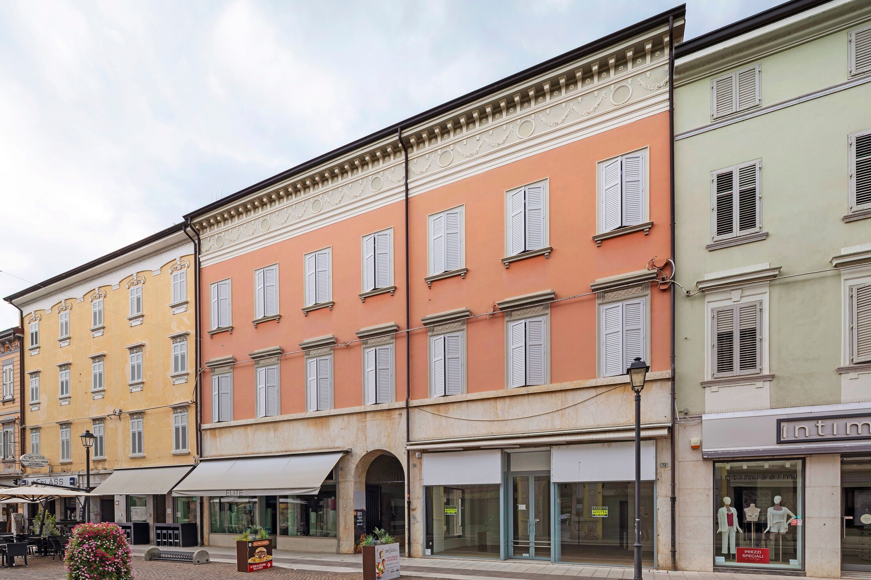 A row of multi-story buildings with uniform window shutters, a street with a café, and a shop with mannequins.