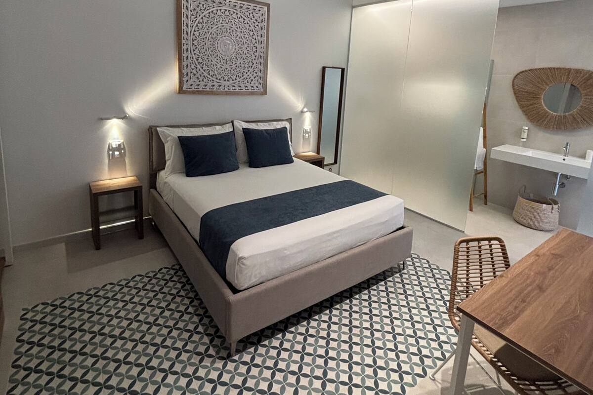 A modern bedroom with a bed, bedside tables, a bedside lamp, a wall-mounted mirror, a small table, and a chair.