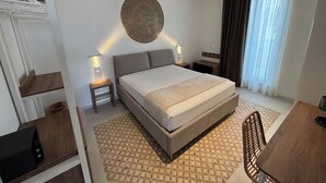 Hotelkamer met een groot bed, nachtkastjes, een decoratief object aan de muur, een raam met gordijnen en een klein tafeltje met een stoel.