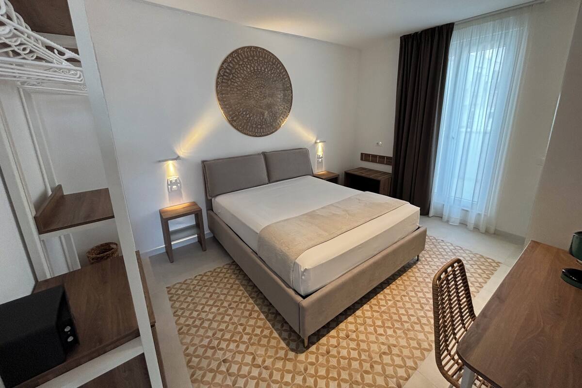A hotel room with a large bed, bedside tables, a wall-mounted decorative piece, a window with curtains, and a small table with a chair.