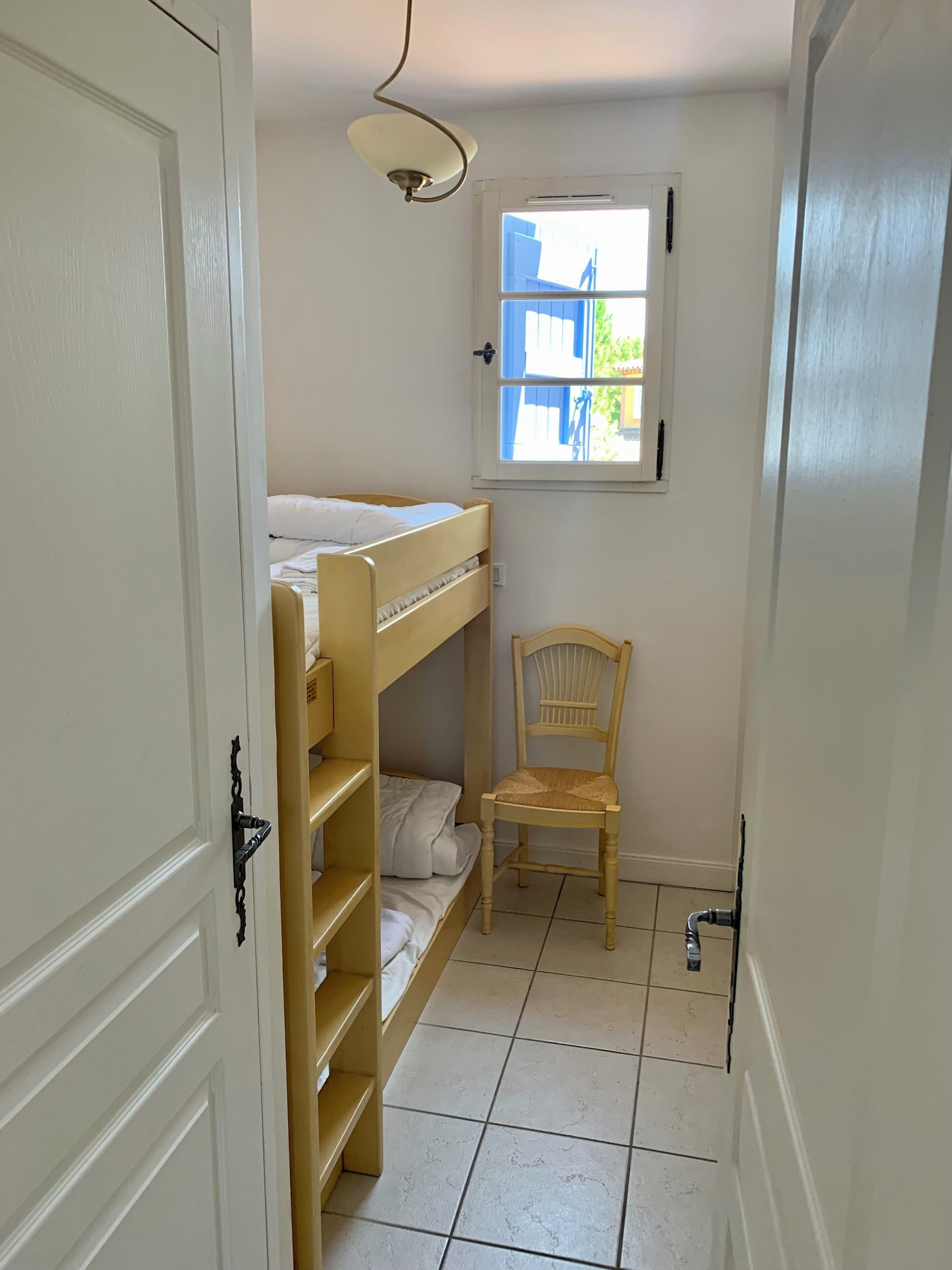 A small, narrow room with a bunk bed, a chair, a window, and a door.