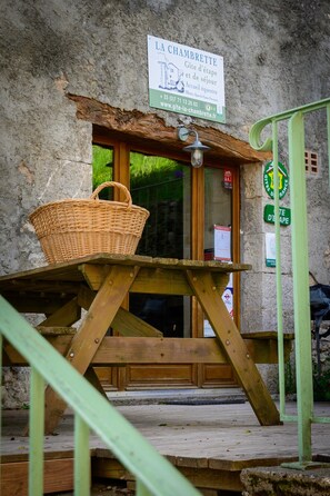 Un banco de madera rústico con una cesta de mimbre, un edificio de piedra con un cartel que dice 'LA CHAMBRETTÉ' y una barandilla de metal verde.