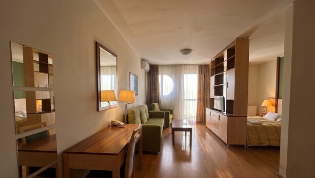 A hotel room with a wooden desk, a green sofa, a small table, a bed with a TV, and a large mirror on the wall.