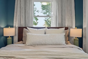 A neatly made bed with a headboard, two bedside lamps, and a window with curtains. - Welcome to Sue Zen Urban Retreat — Grandview Heights, Columbus (Grandview Heights)