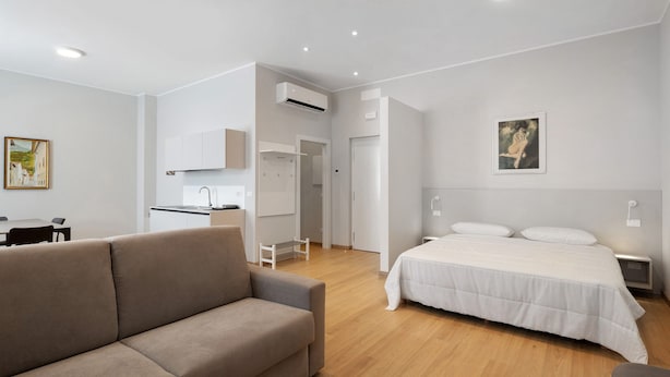 A modern hotel room with a sofa, a bed, a kitchenette, and a painting on the wall.