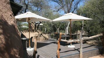 Una terraza de madera con mobiliario exterior y sombrillas, rodeada de árboles.
