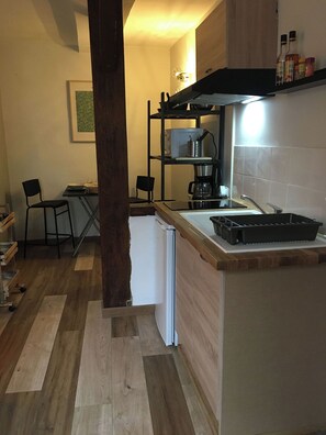 Eine kleine Küche mit einem weißen Kühlschrank, einer Kaffeemaschine und einer Mikrowelle auf einer Ablagefläche. Ein Holzboden und ein Stuhl sind im Hintergrund zu sehen.