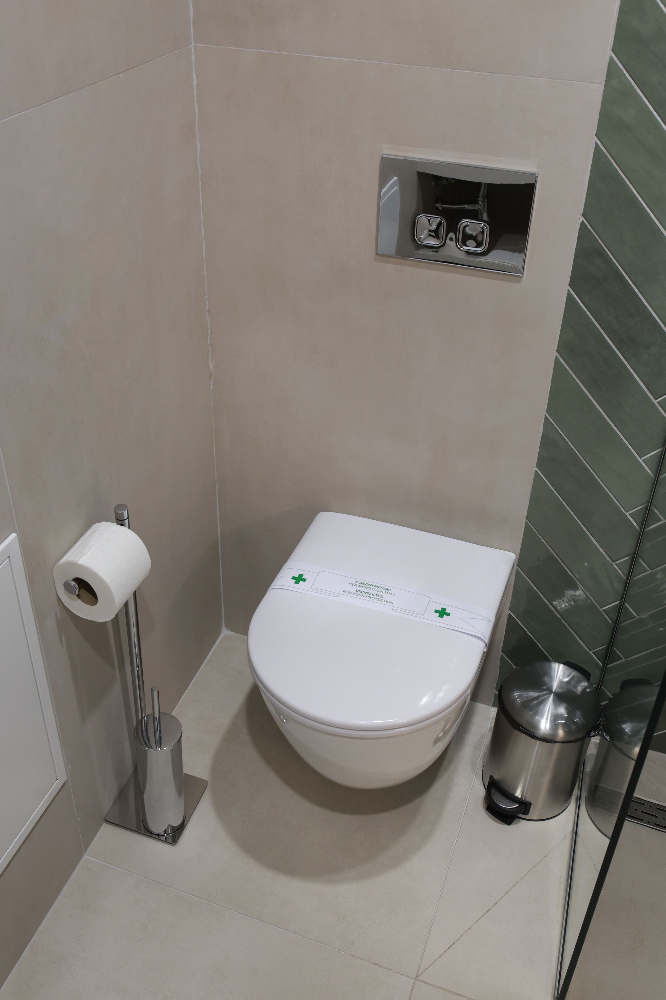 Un baño con inodoro, un dispensador de toallas de papel, un cesto de basura y una pequeña estantería con un par de gafas.