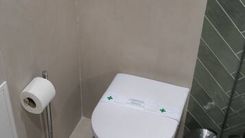 Un baño con inodoro, un dispensador de toallas de papel, un cesto de basura y una pequeña estantería con un par de gafas.