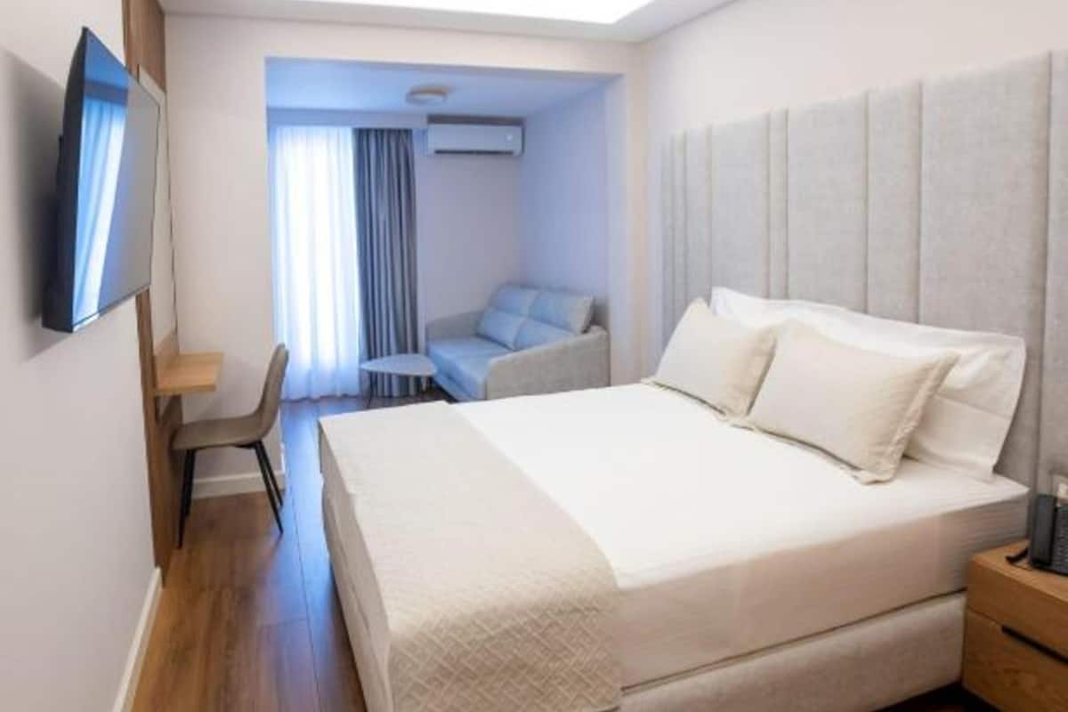 A modern hotel room with a large bed, a desk, a chair, a TV, and a ceiling light.