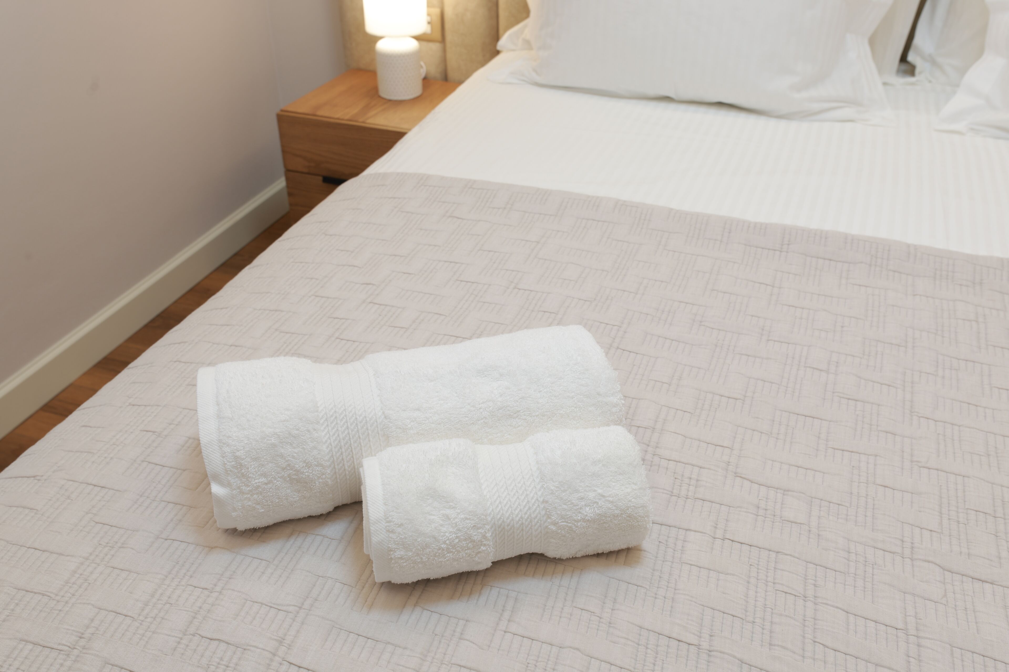 A neatly made bed with a white bedspread and two rolled white towels placed on the bed.