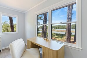 Una habitación con escritorio, silla y ventanas amplias que dan vistas a árboles y una masa de agua.