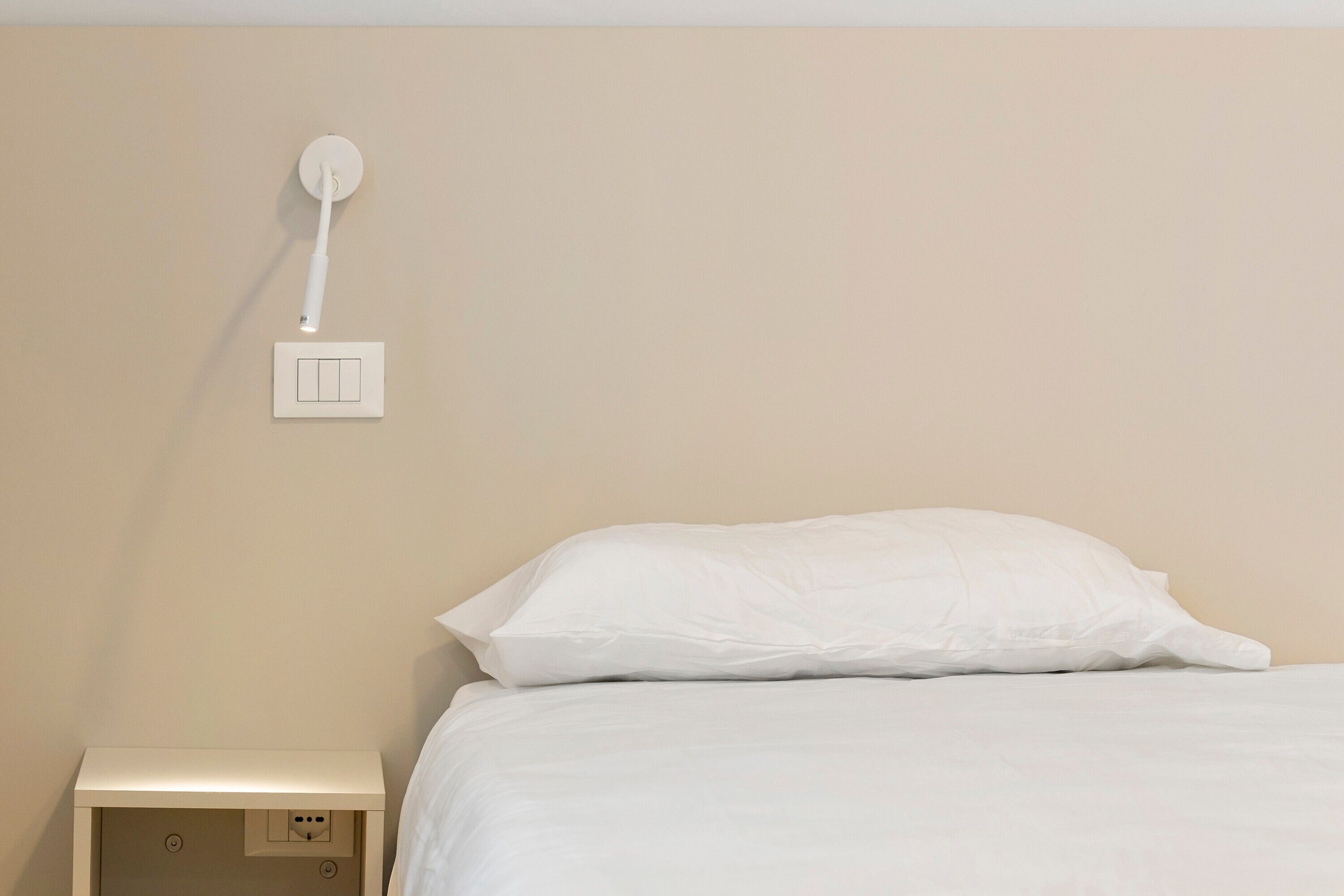 Una cama con ropa blanca, una mesita de noche blanca y una lámpara de pared.