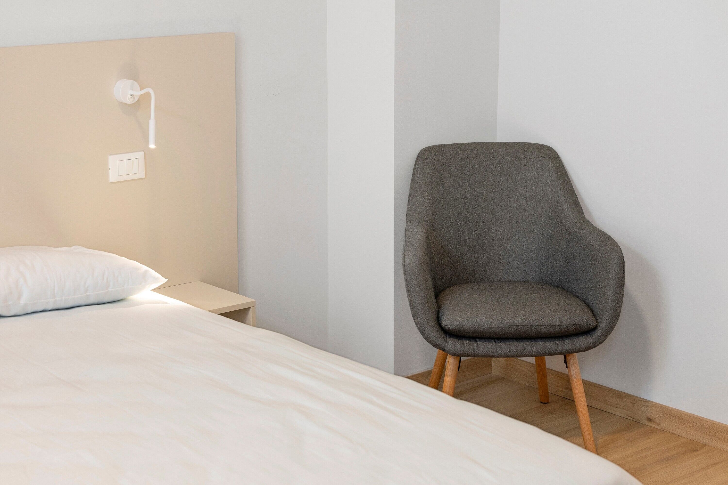 A bedroom with a bed, a nightstand, a chair, and a wall-mounted light fixture.
