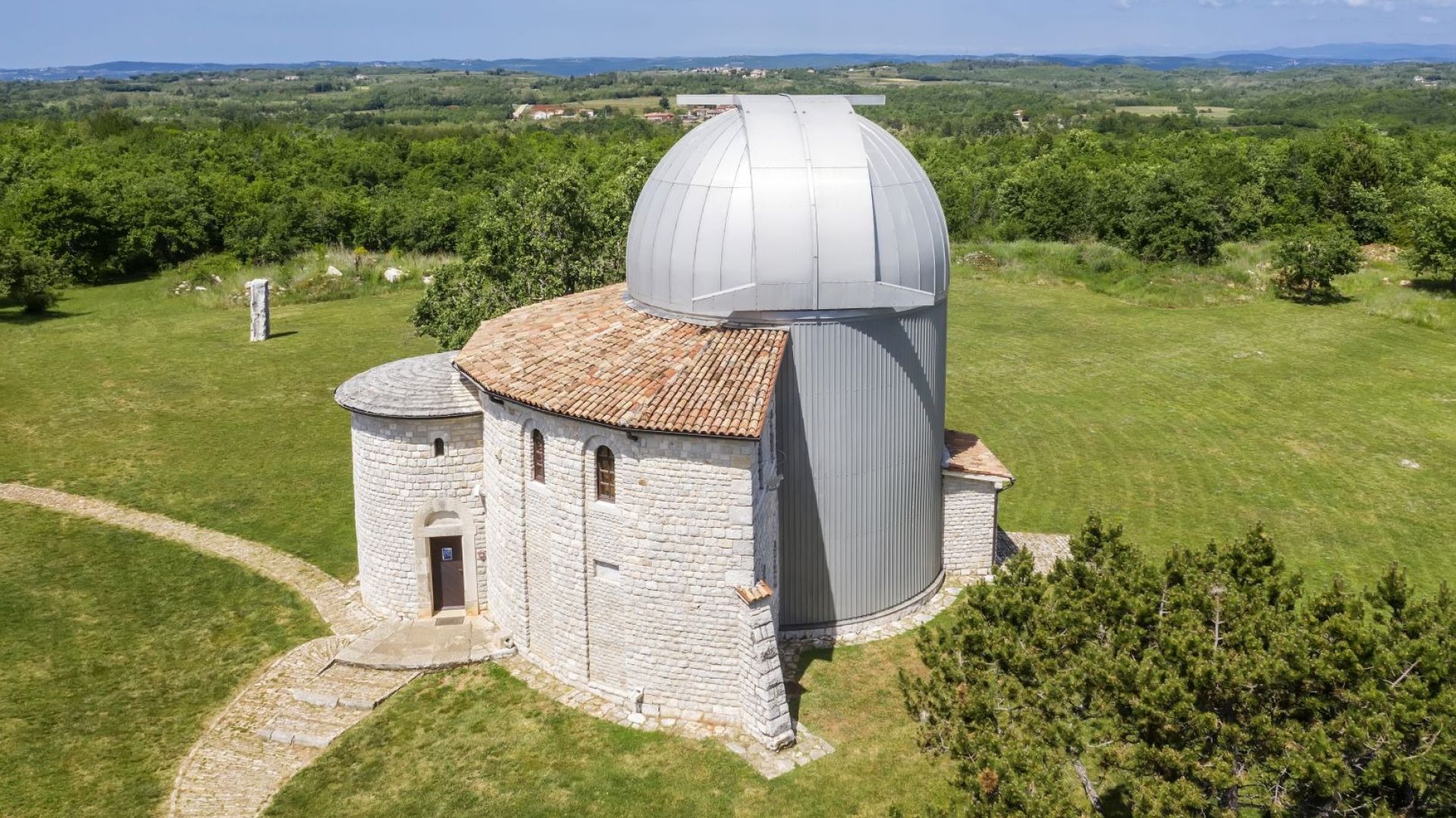 A stone observatory with a dome and a smaller circular building, surrounded by a grassy field and trees.